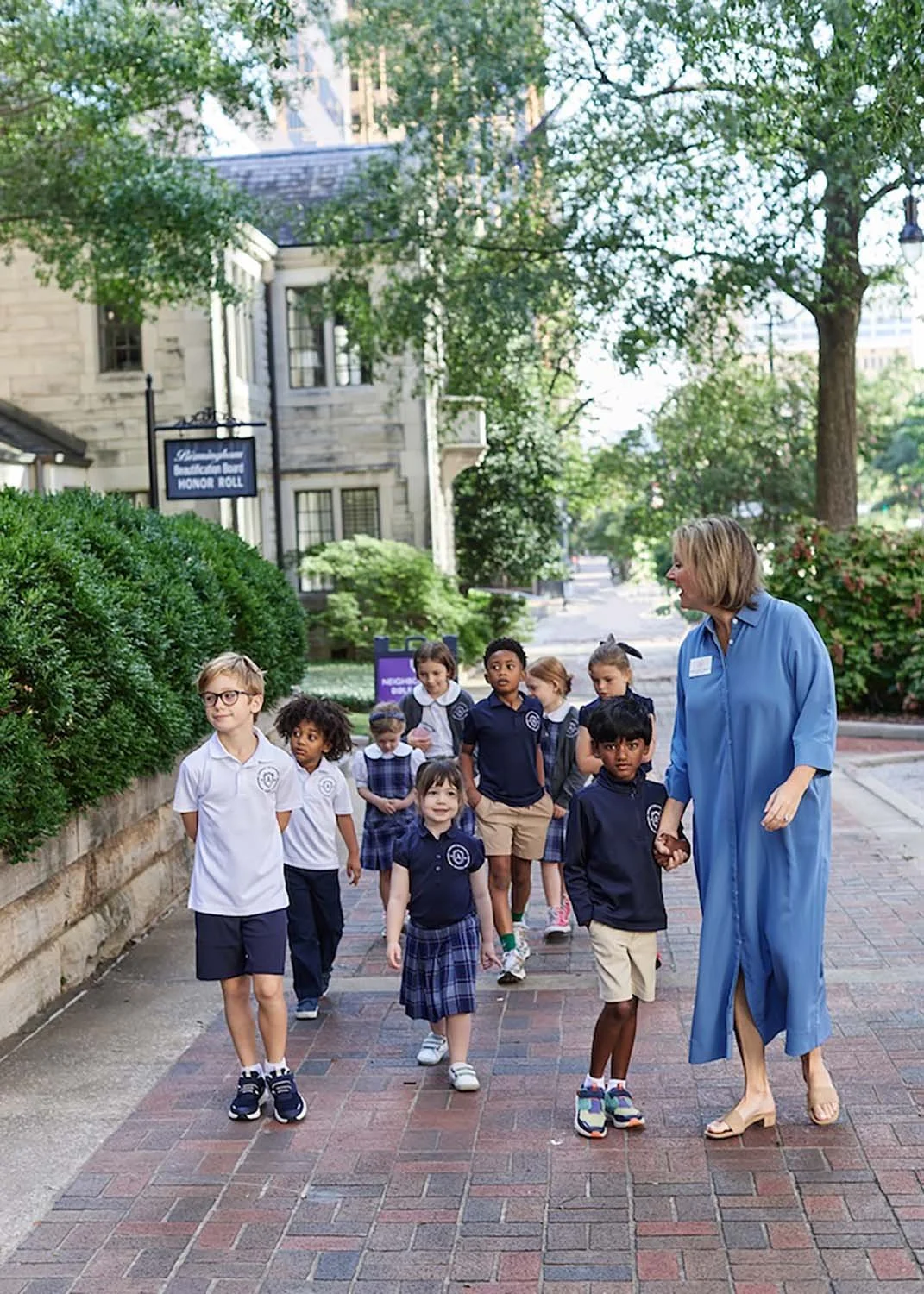 A group of diverse children walking outside on a brick pathway. The children are wearing Advent school uniforms in white and navy blue, with some in plaid skirts and shorts. The area has greenery and the school building in the background.