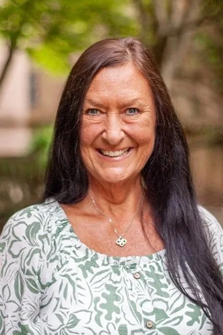 A woman with dark hair and blue eyes smiling outdoors with green foliage in the background.