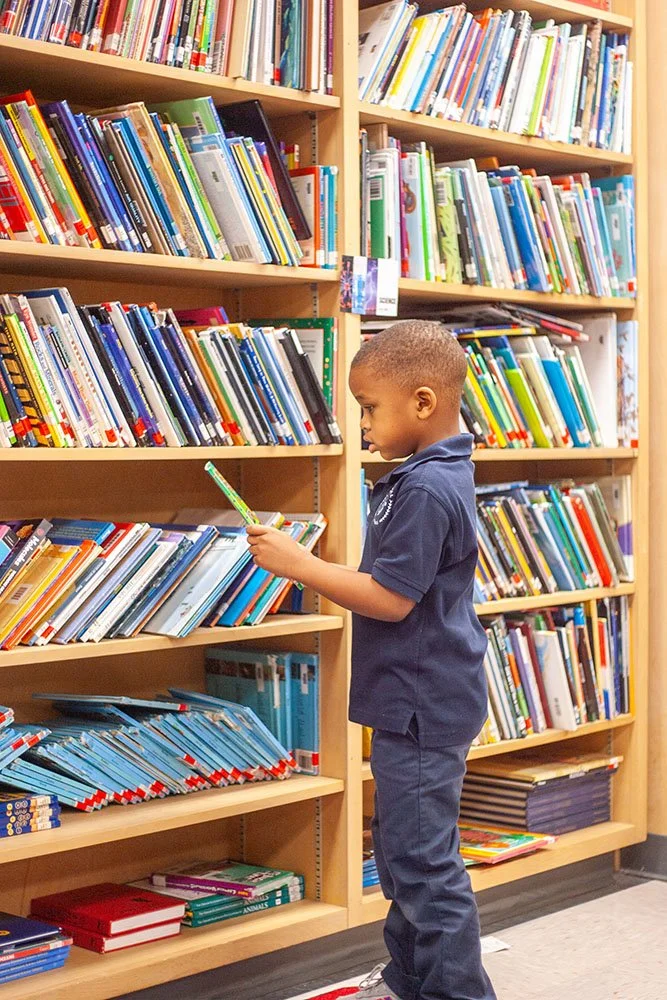 A young boy standing in a library browsing through a book on a wooden bookshelf filled with colorful books.