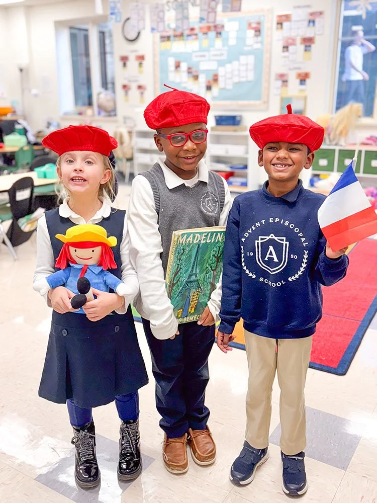 Three children wearing red berets in a classroom, holding French-themed items, smiling and standing together.