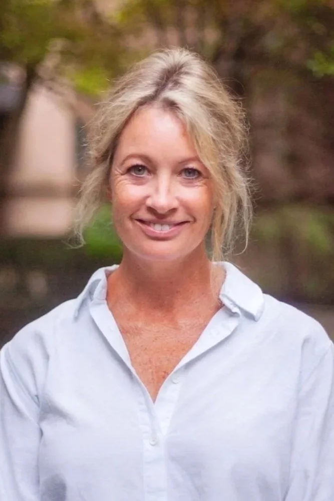 A woman with blonde hair, wearing a white textured top, smiling outdoors with greenery in the background.