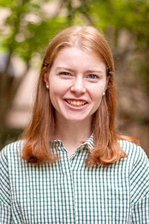 A young woman with red hair, freckles, and blue eyes smiling outdoors in a light green shirt with a blurred green leafy background.