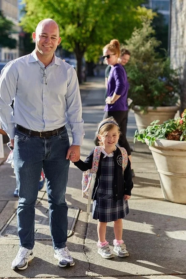 A man and a young girl holding hands outside on a sidewalk, with other people and large planters with flowers in the background.