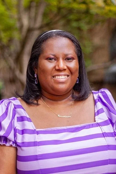 A woman outdoors wearing a purple and white striped dress, with a necklace and earrings, smiling at the camera.