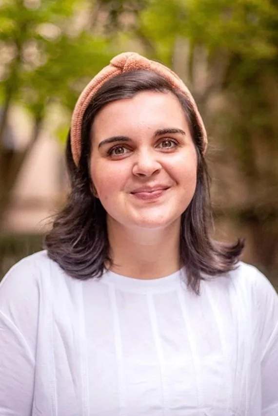 A young woman with dark hair and brown eyes smiling, wearing a white shirt, standing outdoors with green bushes and fallen branches in the background.