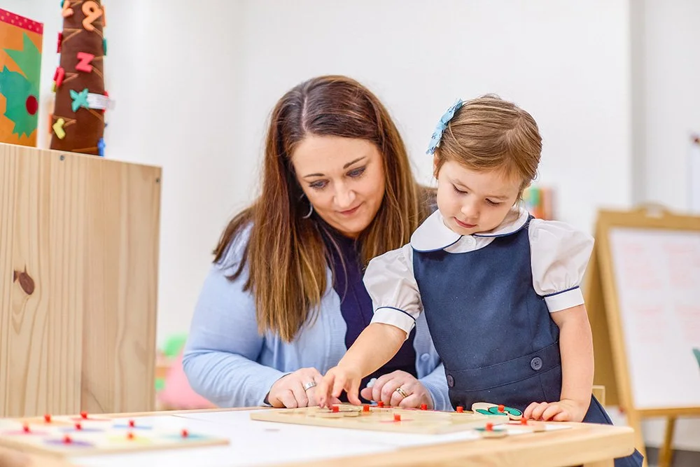 A teacher woman and a young girl playing a board game together at a table in a classroom or playroom.