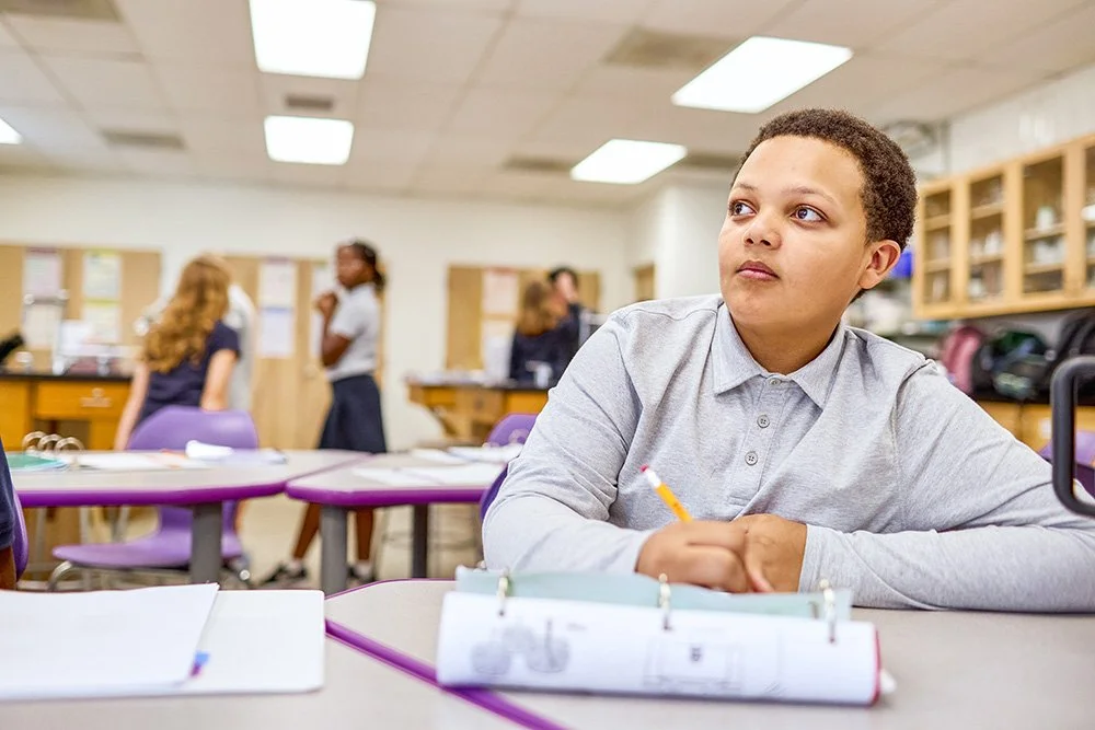 A young boy sitting at a classroom desk with a pencil, looking to the side. In the background, several girls are standing and chatting in the classroom.
