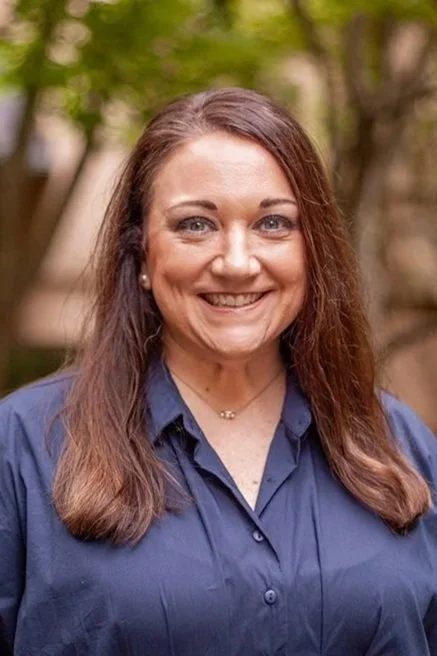 A woman with long brown hair, blue eyes, wearing a gray top and earrings, smiling outdoors with green foliage in the background.