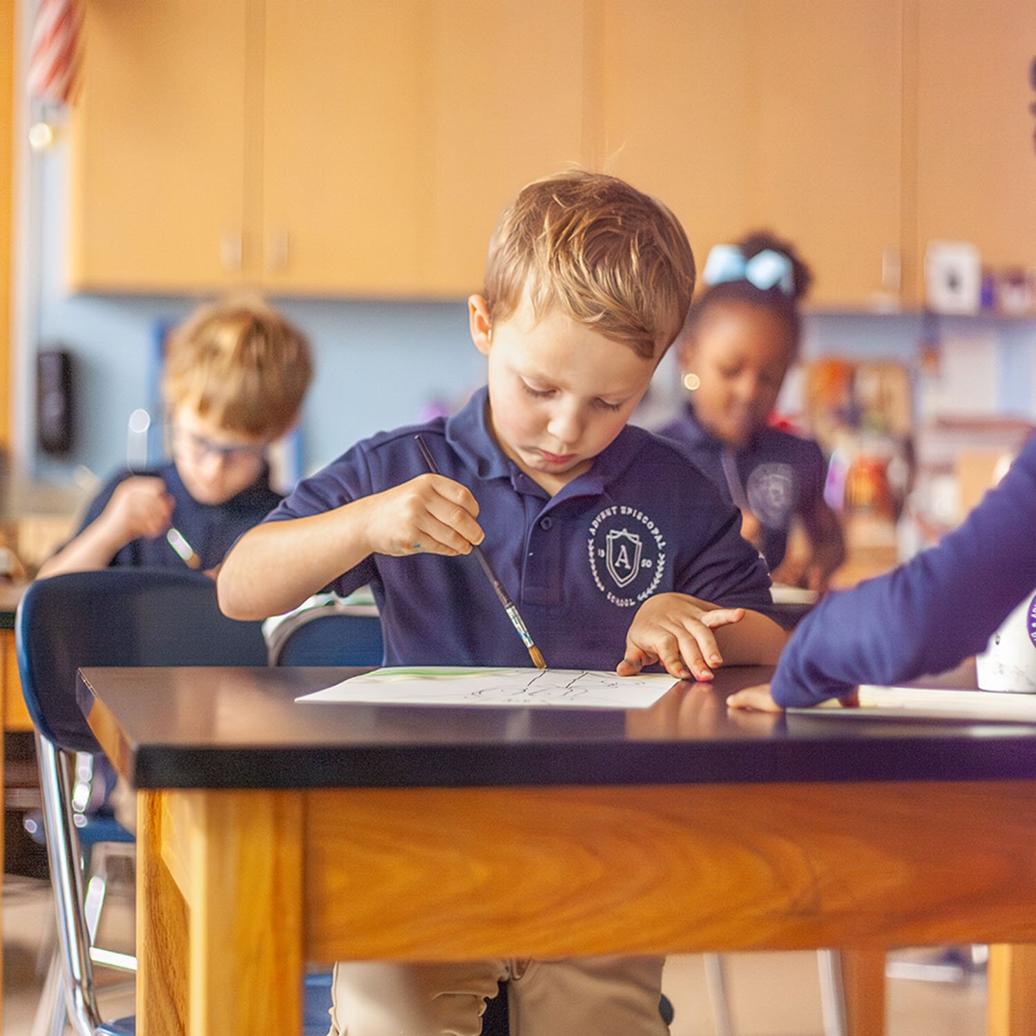 A young boy with light brown hair, wearing a navy blue school uniform shirt, is sitting at a desk drawing with a paintbrush on paper. There are other children in the background also working at their desks in a classroom setting.
