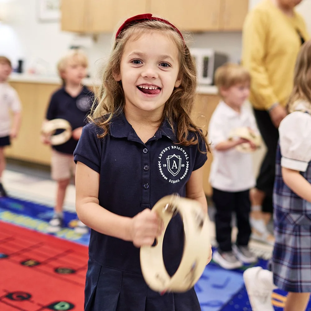 A smiling young girl with long curly hair wearing a navy blue school uniform and red headband, holding a tambourine in a classroom with other children and an adult in the background.