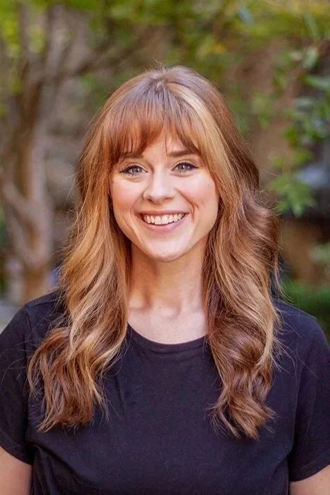 A smiling woman with shoulder-length brown hair and bangs, wearing a black button-up shirt, standing outdoors with greenery in the background.