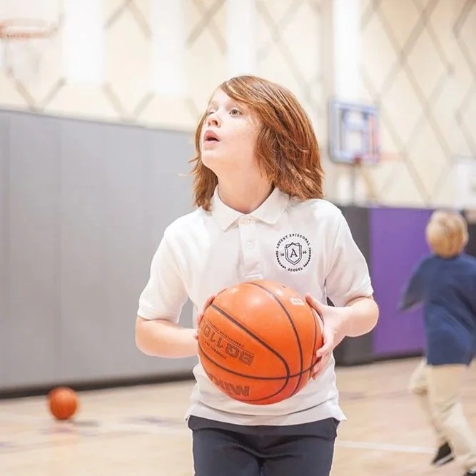 A young boy with red hair holding a basketball inside a gymnasium, with a boy running in the background.