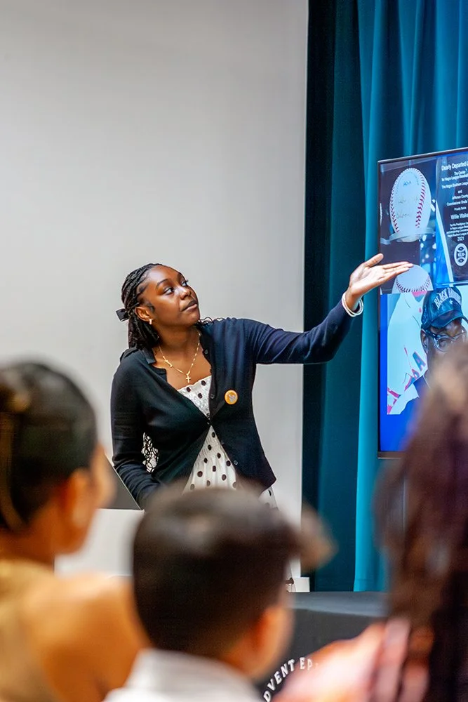 A student giving a presentation or lecture to a group of people, standing next to a large screen displaying graphics, with an audience in the foreground.