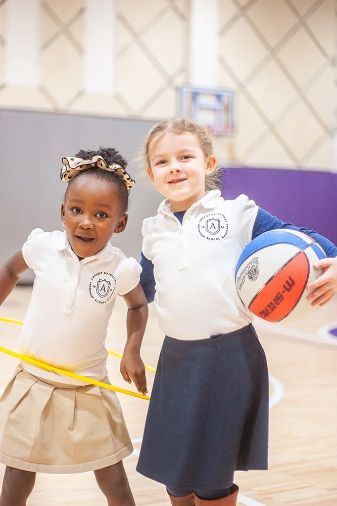 Two young girls in school uniforms participating in a gym class; one girl holds a basketball, both are smiling.