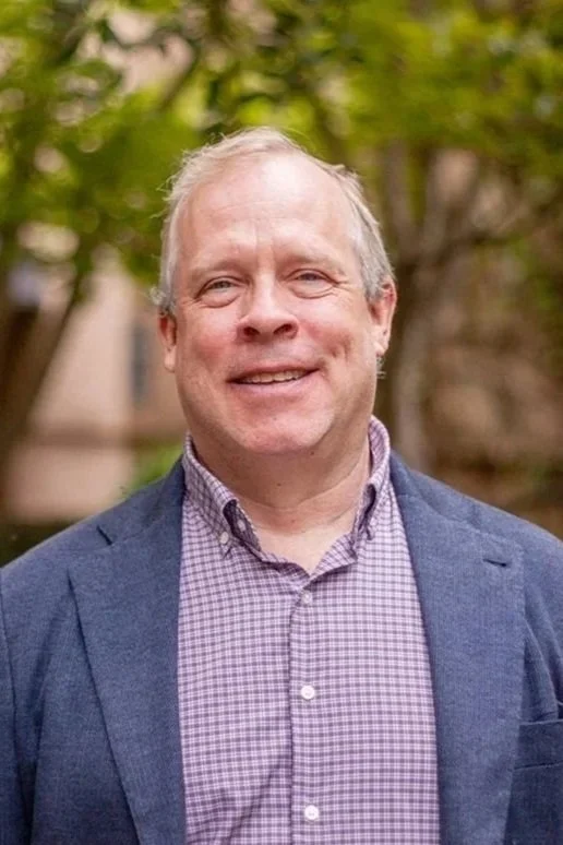 Smiling middle-aged man in a striped collared shirt standing outdoors in front of green foliage.
