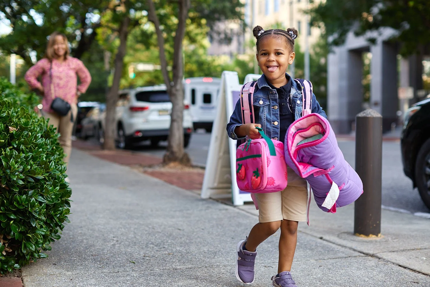 A young girl with pigtails, wearing a denim jacket and shorts, smiling and walking on a city sidewalk while holding a pink lunchbox and a purple jacket. An adult woman is in the background, smiling, near parked cars and trees.