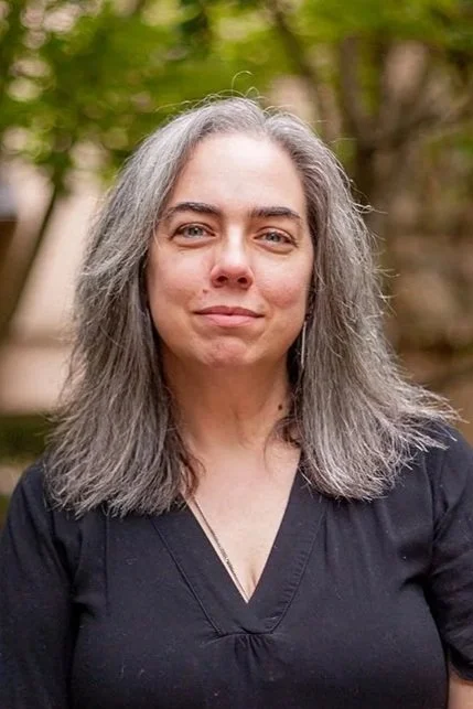 A woman with glasses and long gray hair standing outdoors with greenery in the background.