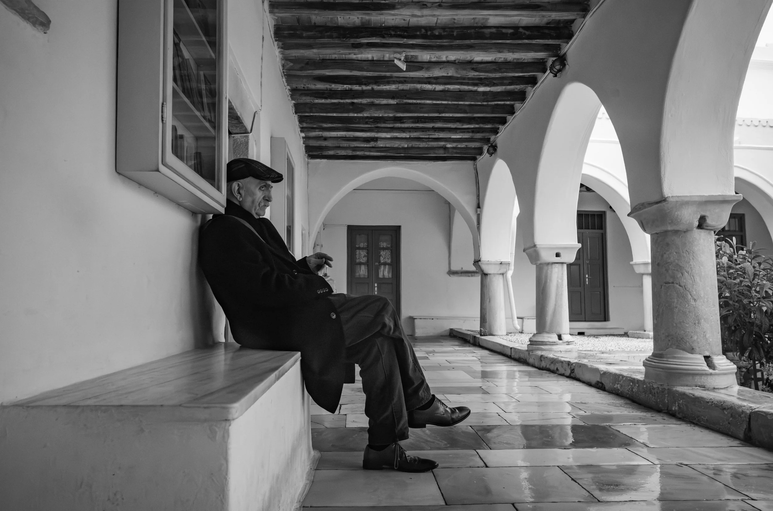 An elderly man wearing a cap, dark coat, and dark pants is sitting on a bench in a covered outdoor corridor with white arches and columns, holding a small object in his hand, next to a window with glass panes.