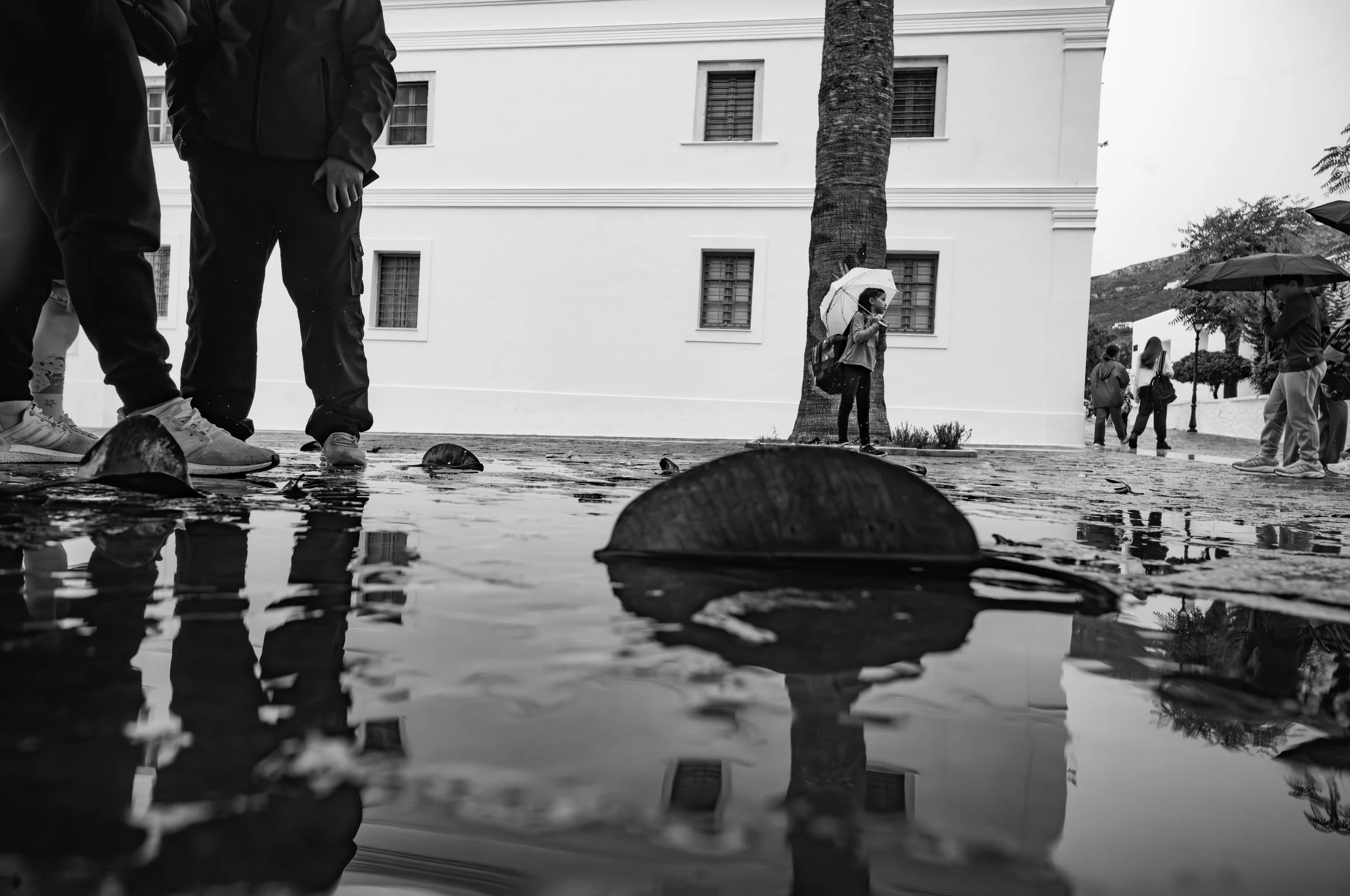 A black and white photo of children standing outside near a building and a tree in the rain, holding umbrellas, with water reflecting objects on the ground.