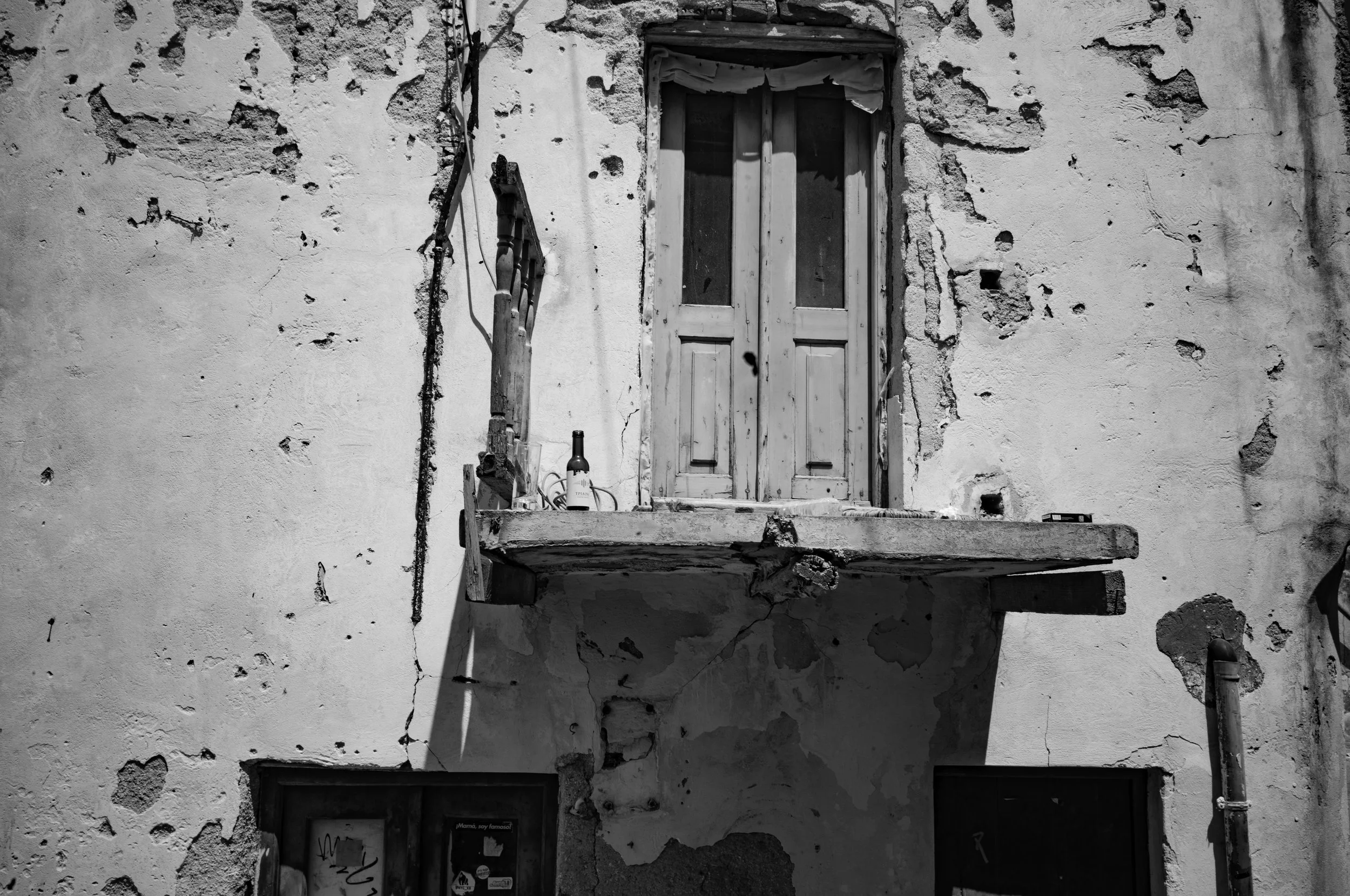 An old, weathered building with a rough, peeling wall and a small wooden balcony with a closed door, a bottle, and some wires hanging nearby, all in black and white.