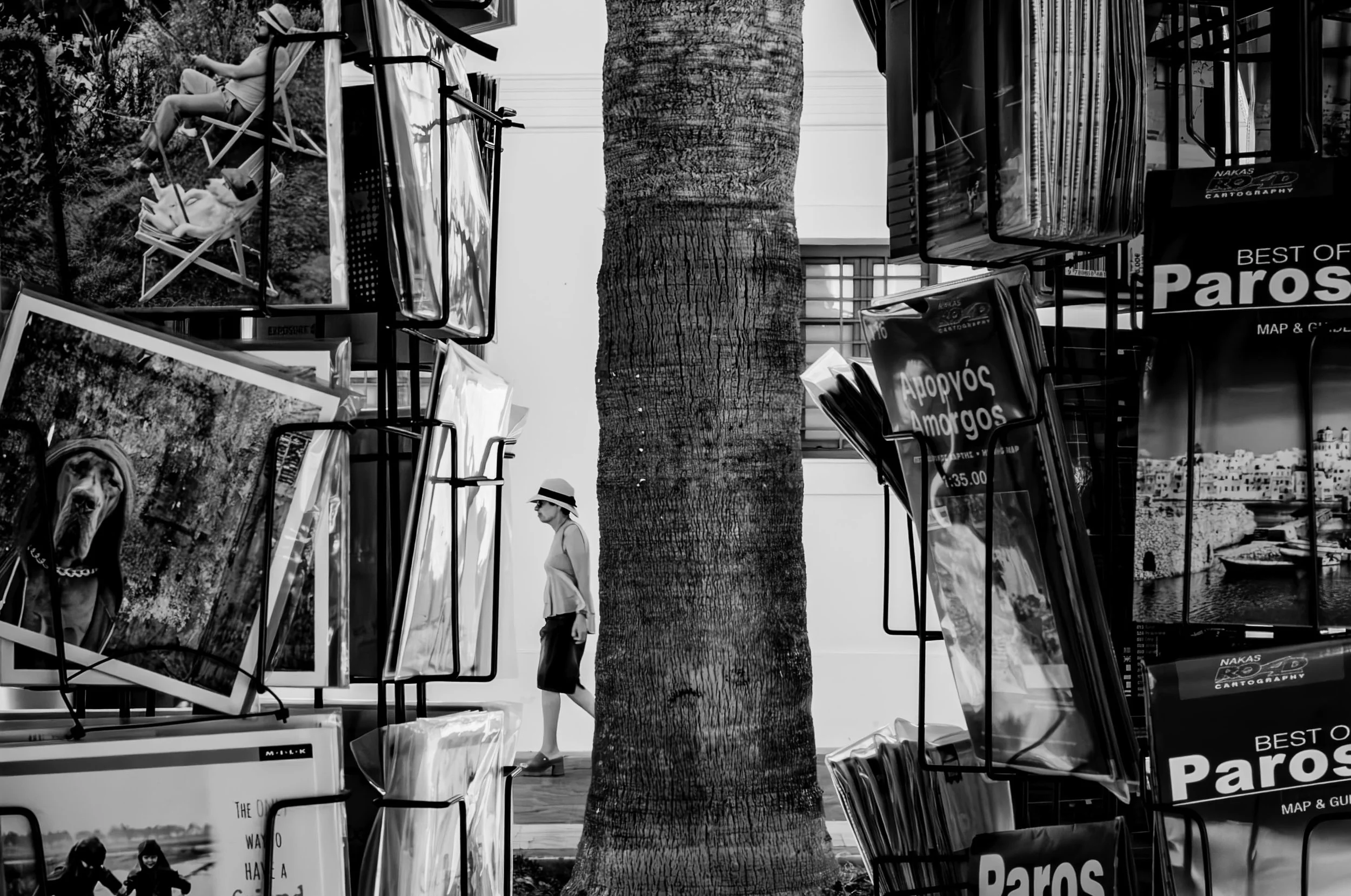 Black and white photo of a woman in a hat and sunglasses walking along a city sidewalk lined with street vendor stalls, a large tree in the center, and a building with windows in the background.