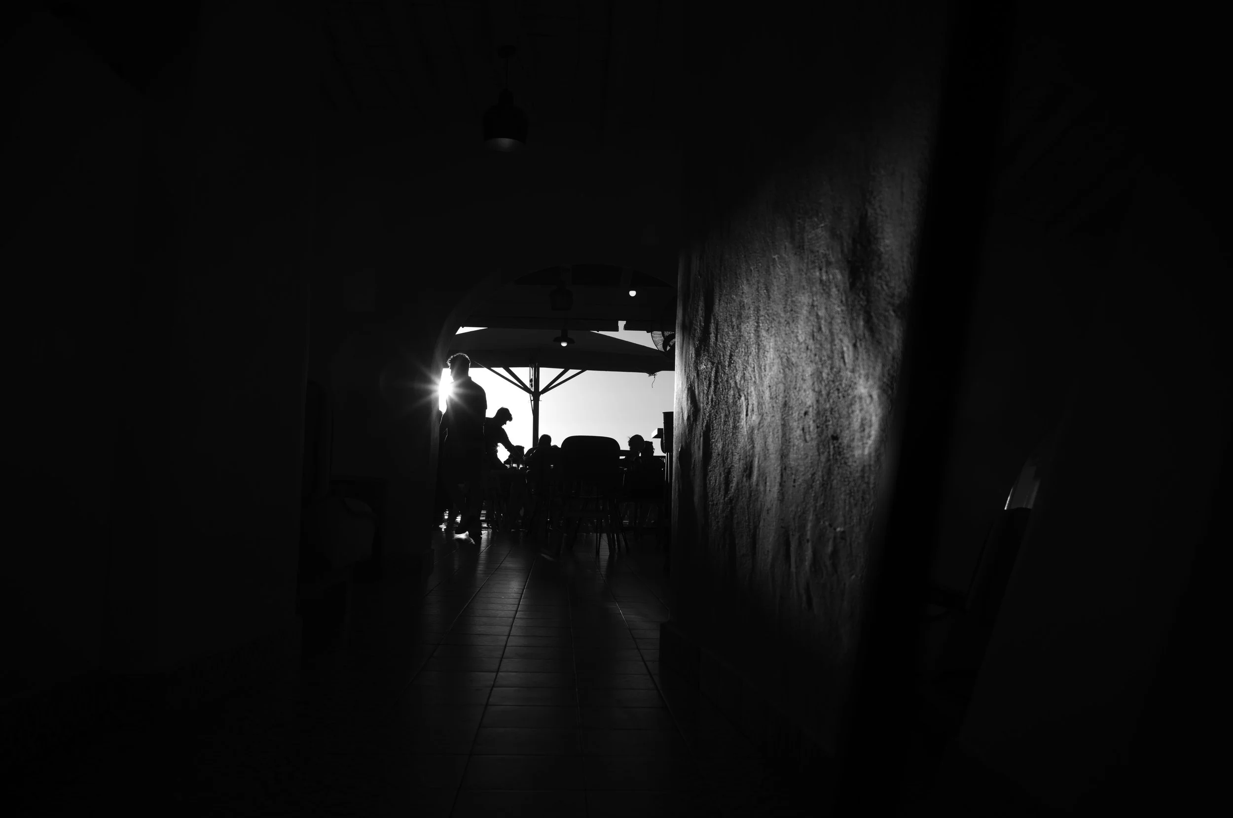 Silhouettes of people inside a dimly lit room with bright sunlight streaming through a window or opening, creating a contrast between dark and bright areas.