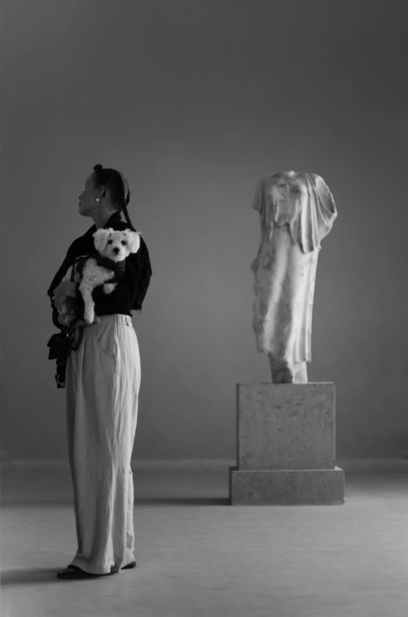 A woman with braided hair holding a small white dog in a gallery with a marble sculpture on a platform in the background.