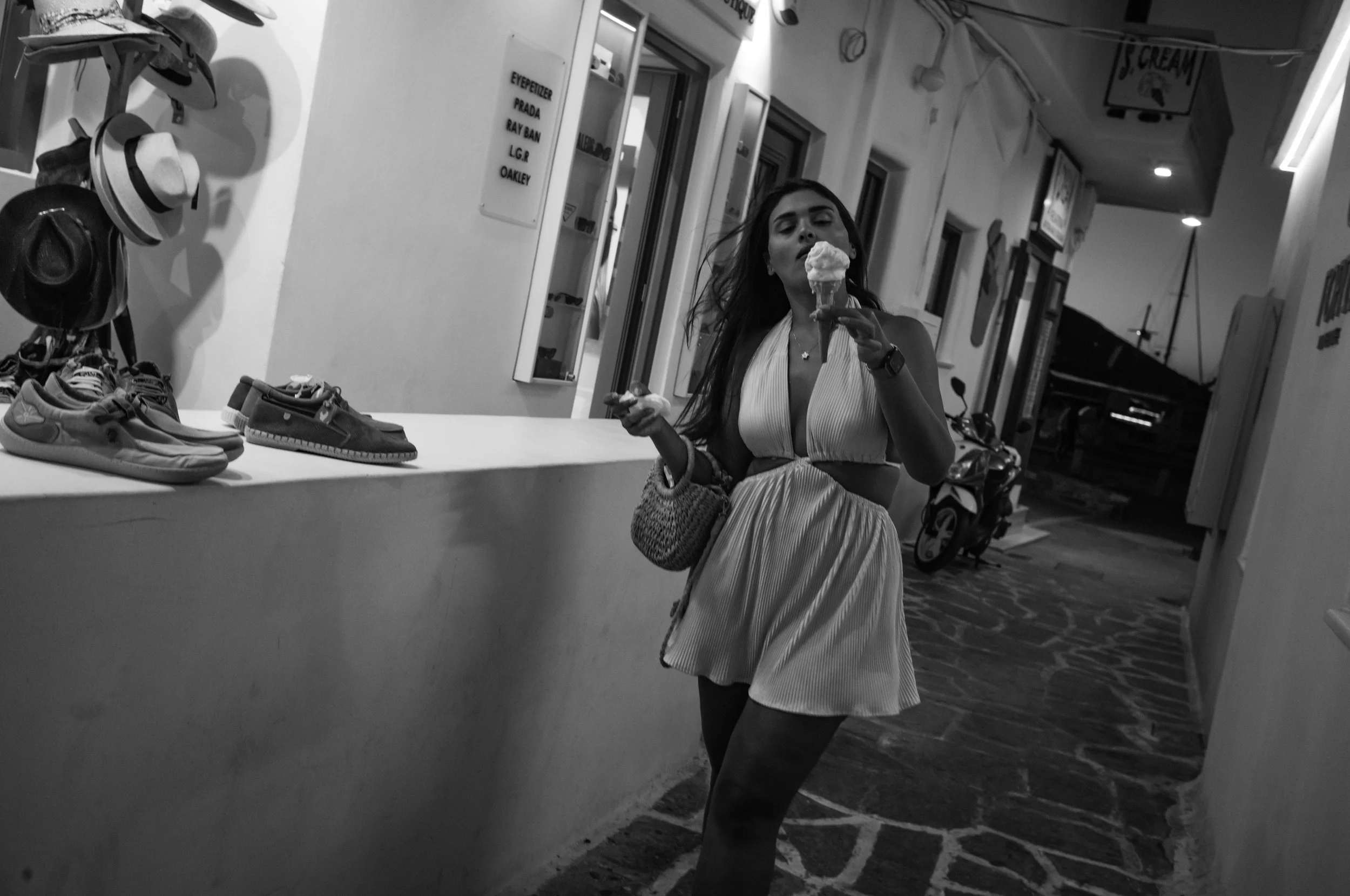 A woman in a sleeveless top and skirt is holding an ice cream cone and a phone, standing inside a shoe store. Shoes are displayed on a counter to her left, and hats hang on the wall behind her. There are signs on the wall with brand names, and the st
