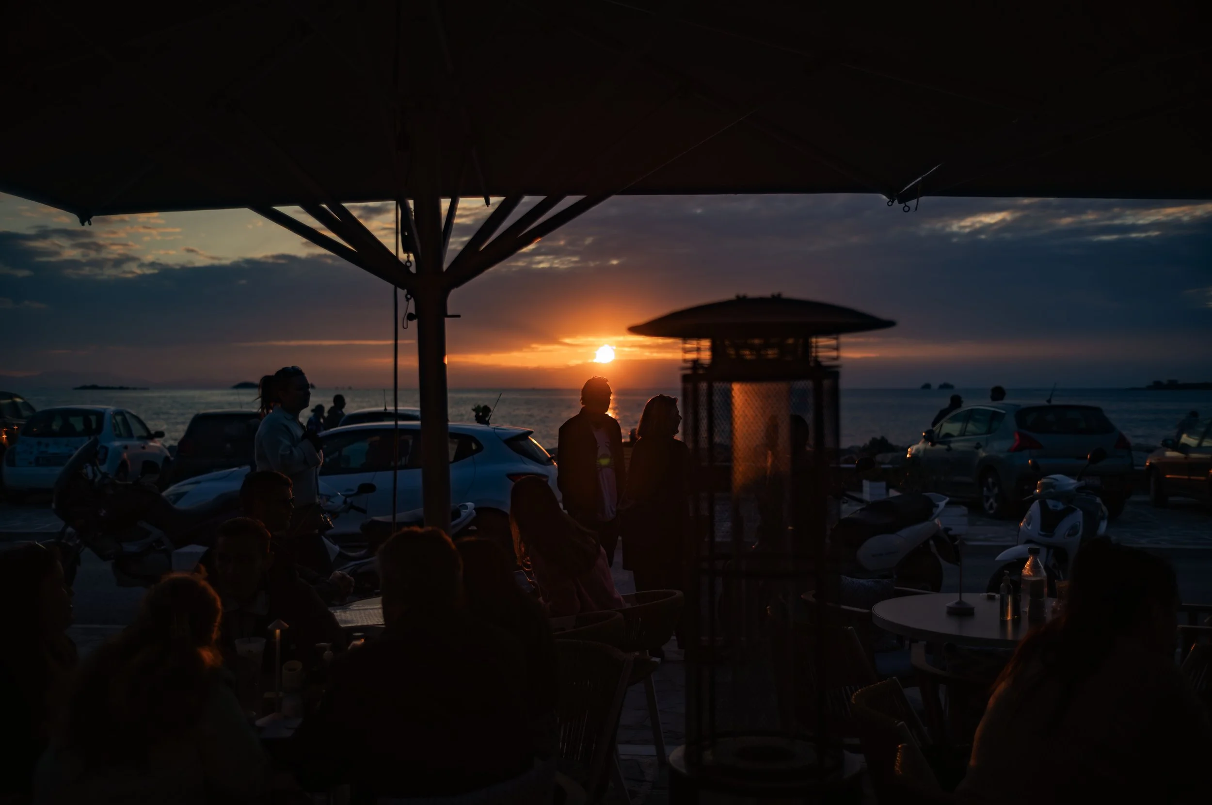 People gathered at outdoor seating area during sunset by the ocean, with cars parked nearby and umbrellas overhead.