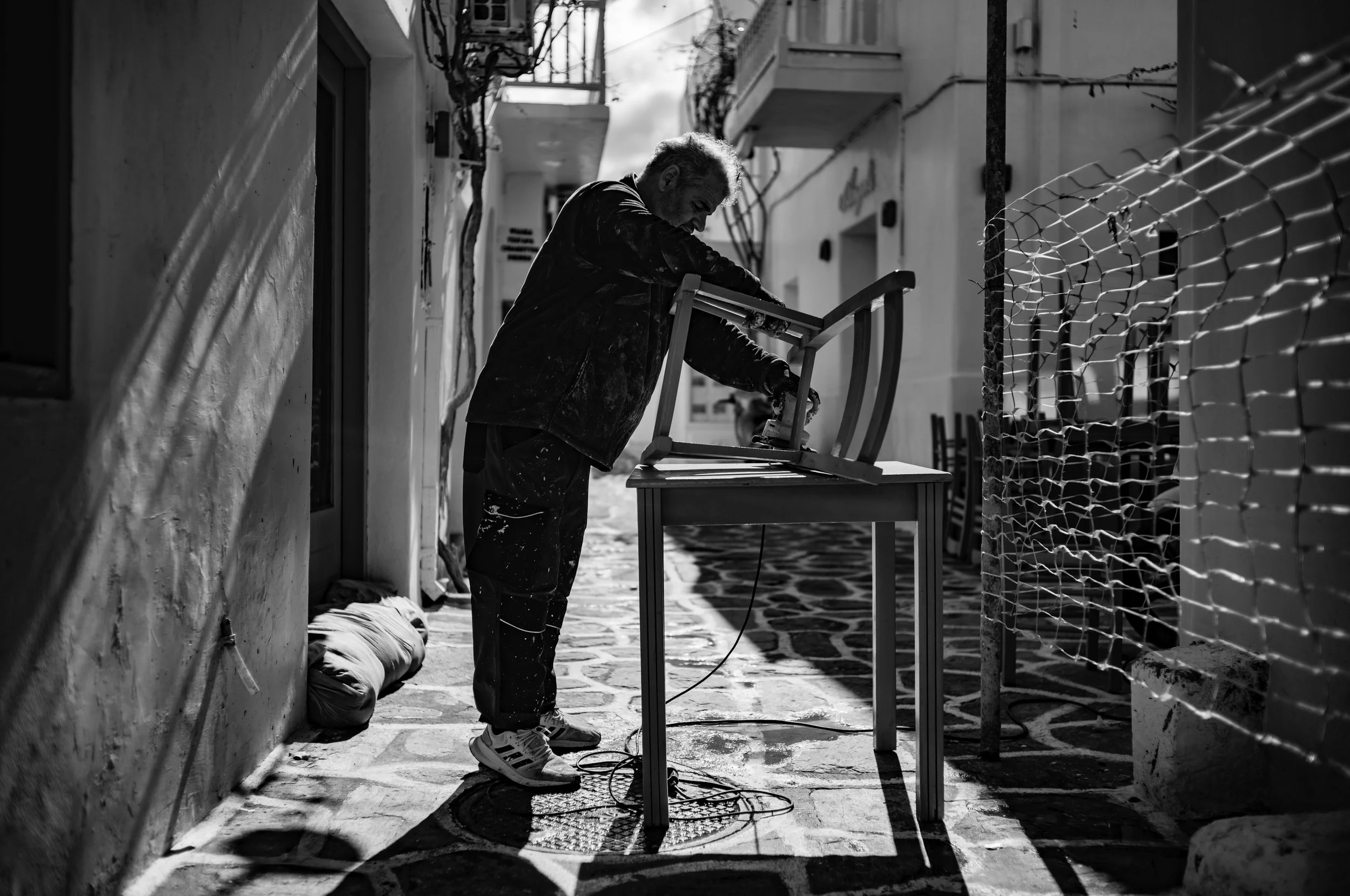 A man working outdoors on a sidewalk, painting a wooden chair. The scene is in black and white, with sunlight casting shadows on the wall behind him.