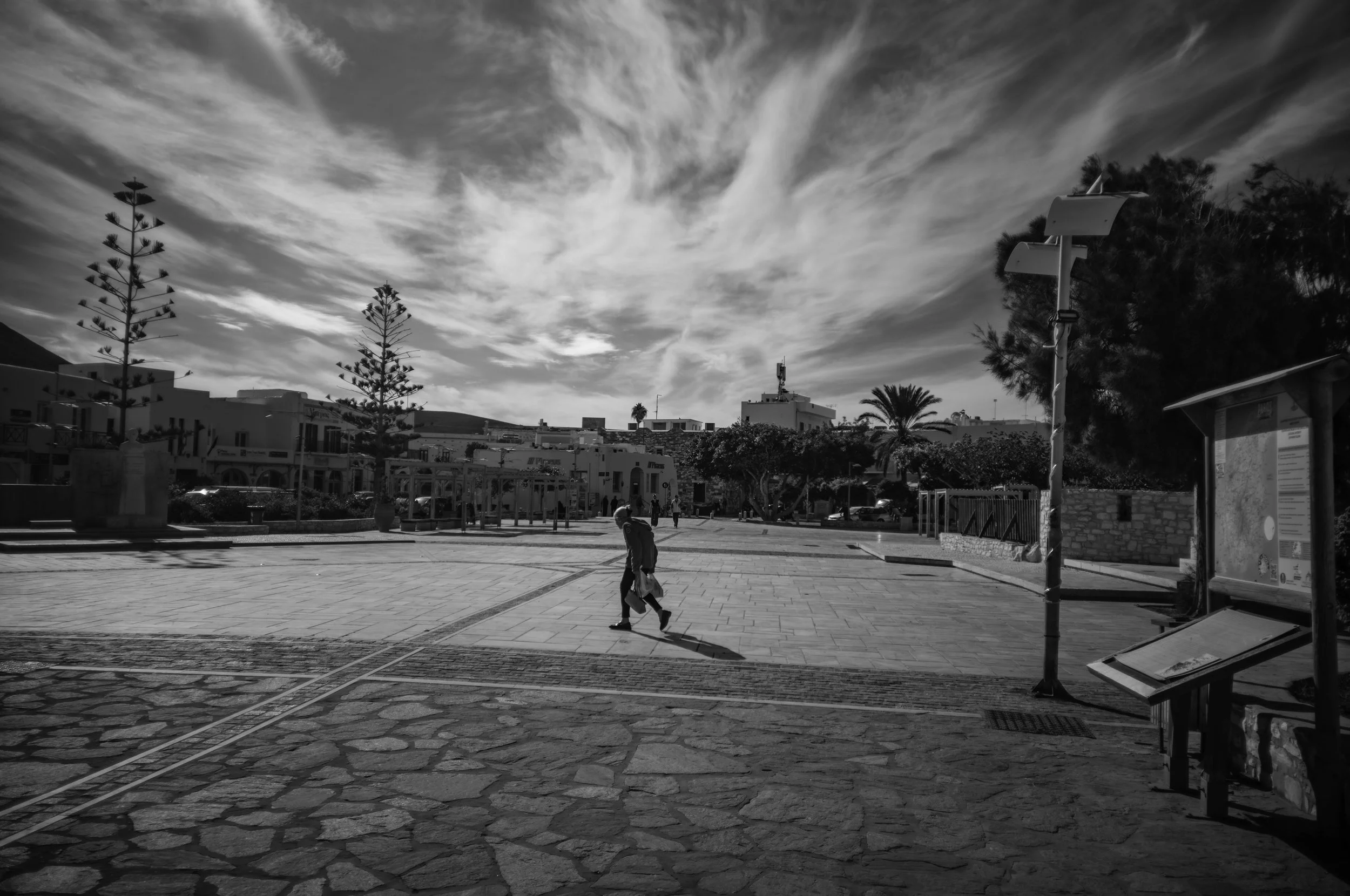 A black and white photo of a town square with a clear sky overhead. There are trees, buildings, and a few people walking across the square. A lamppost with signs or boards is visible on the right side of the image.