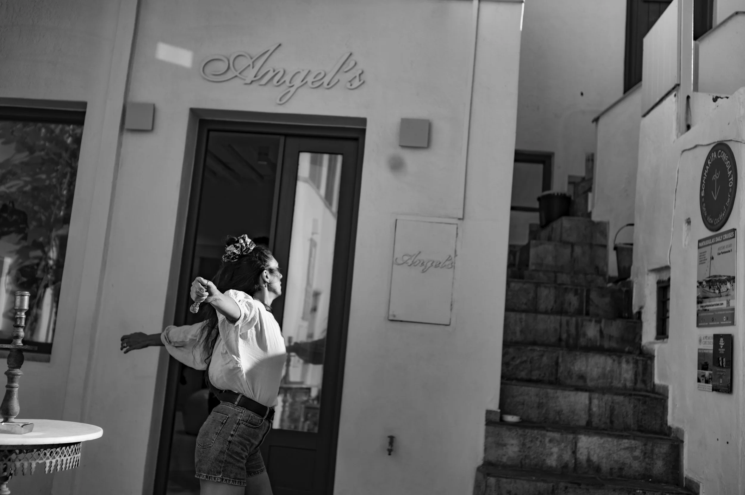 A woman standing outside a building with a sign that reads 'Angel's' on the wall, stretching her arms behind her head. She is wearing glasses, a white shirt, and high-waisted jeans.
