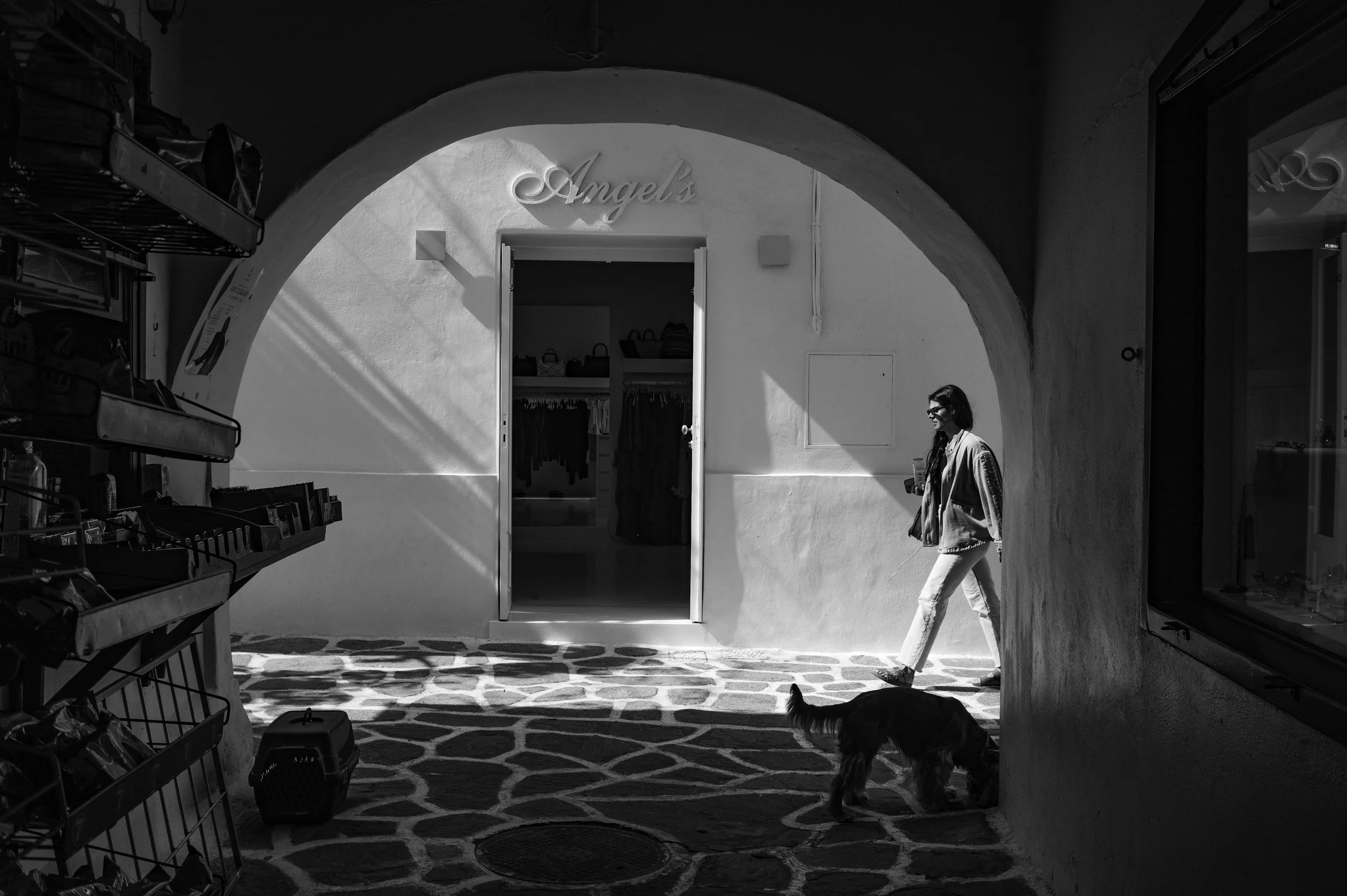 Black and white photo of a shopping area with an arched entrance labeled 'Angel's'. A woman with long hair, sunglasses, and casual clothing walks past a dog near the sidewalk. Store shelves with various items are visible on the left, and the floor ha