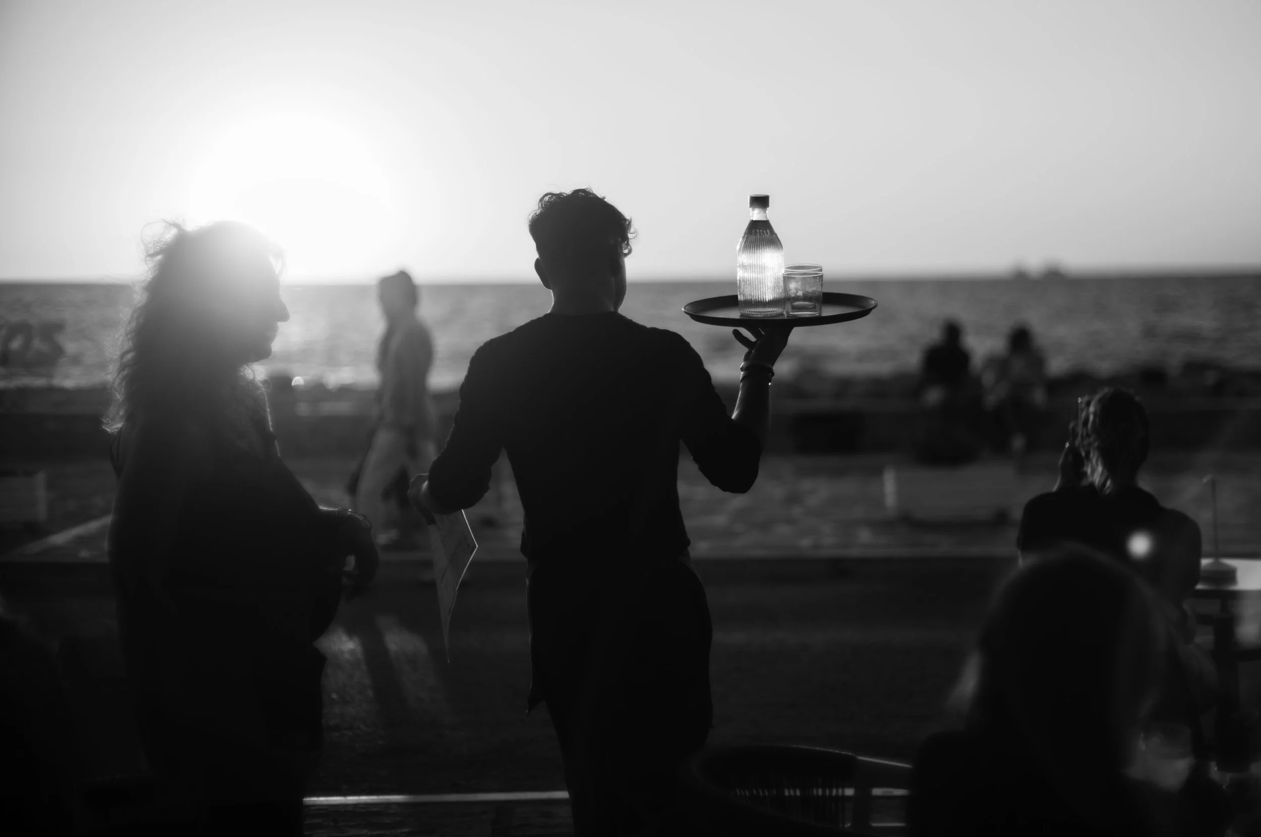 Silhouettes of people at the beach during sunset, with a waiter serving drinks in the foreground.