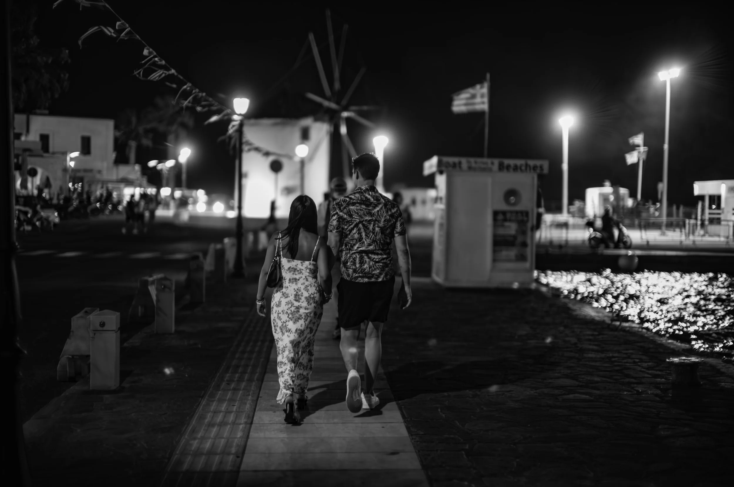 A couple with a woman in a floral dress and a man in a patterned shirt walking down a well-lit night promenade, with a windmill sculpture and flags in the background.