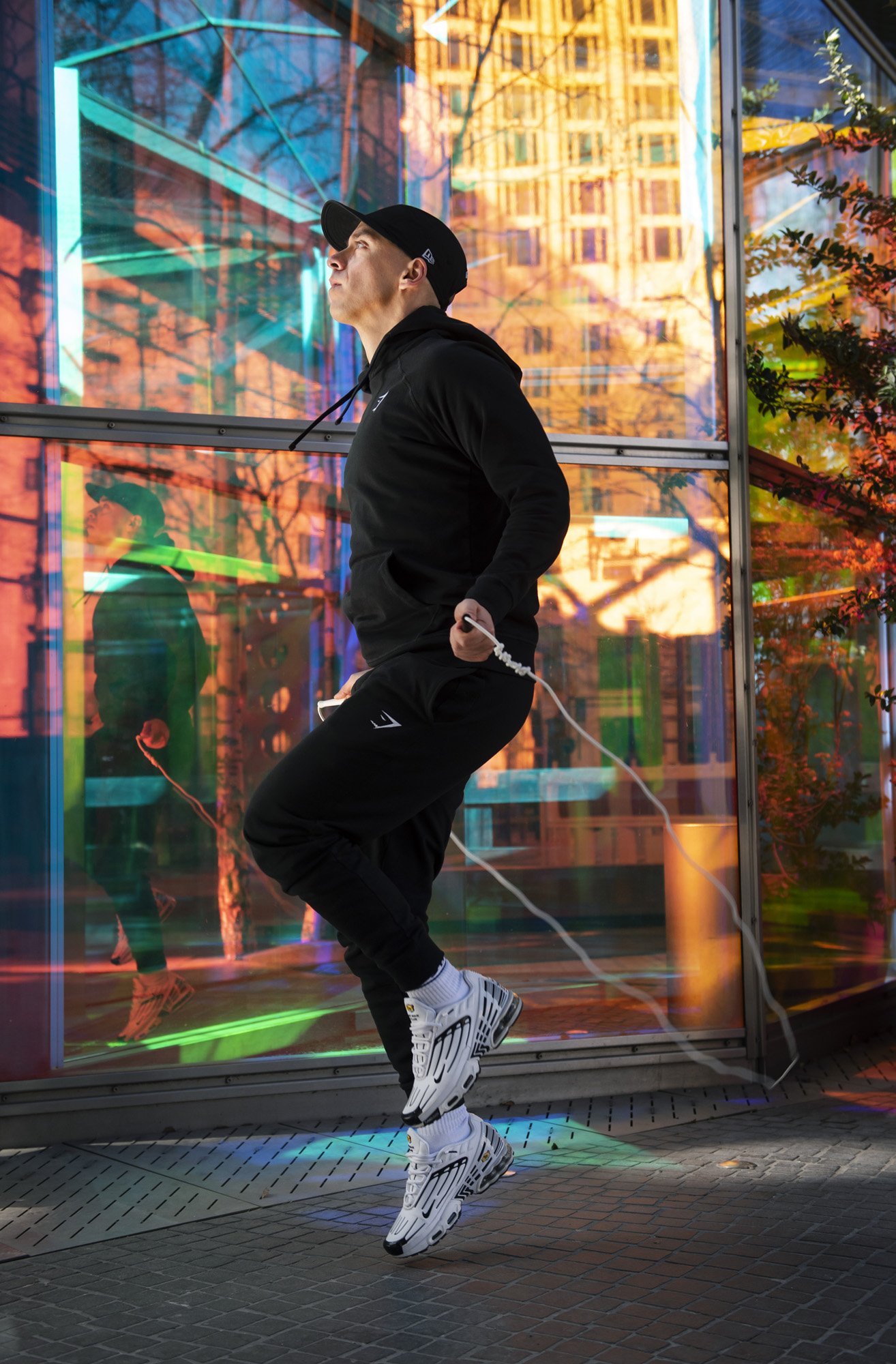 Person in black athletic wear and cap jumping rope in front of colorful glass building facade.