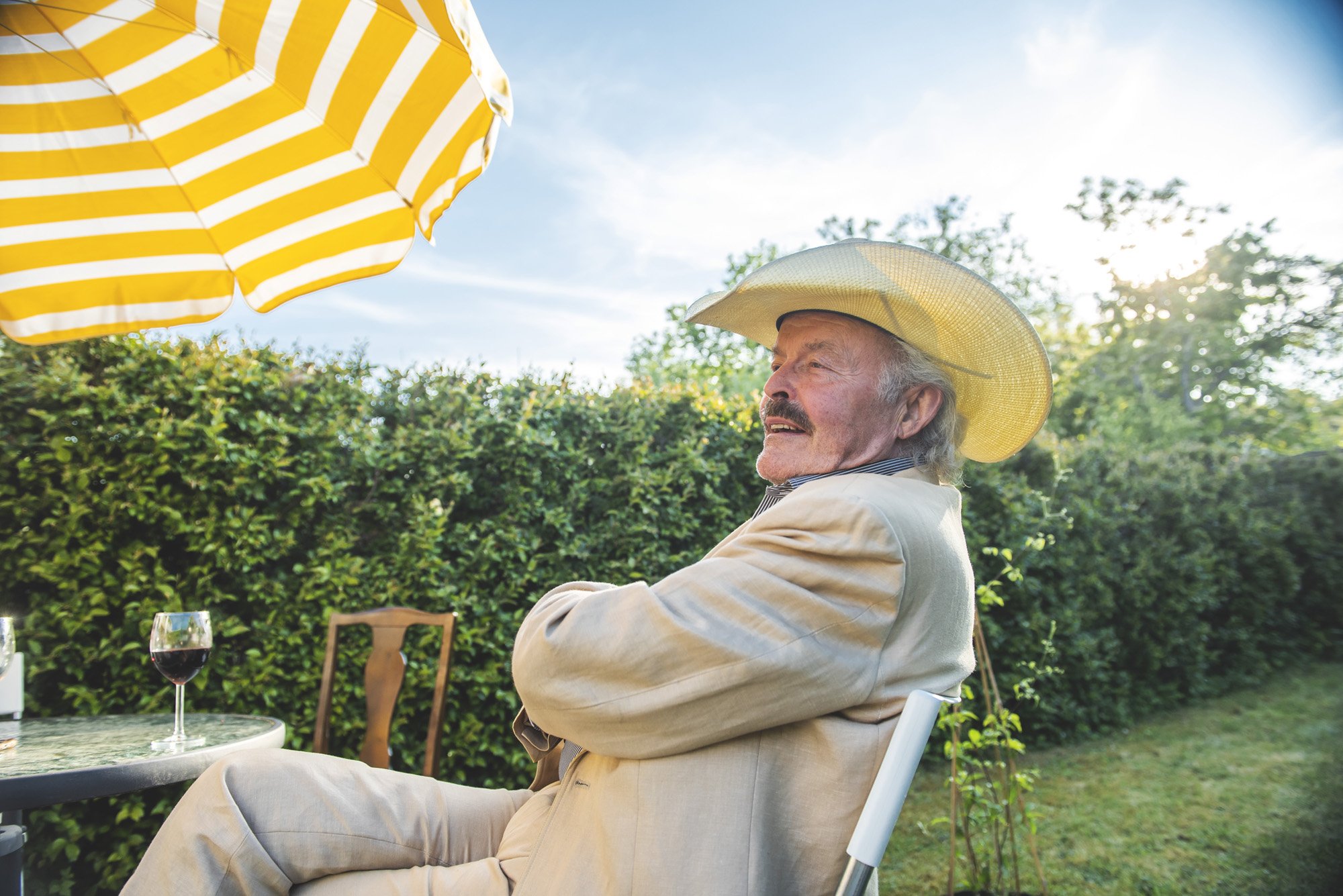 Elderly man wearing a straw hat sitting outdoors in a garden, under a striped yellow umbrella, with a glass of red wine on a table.