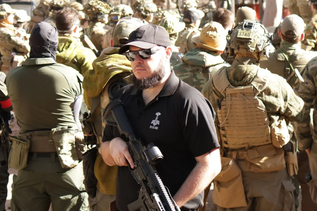 A person wearing a mask, sunglasses, and tactical gear, aiming a firearm with smoke in a wooded area.