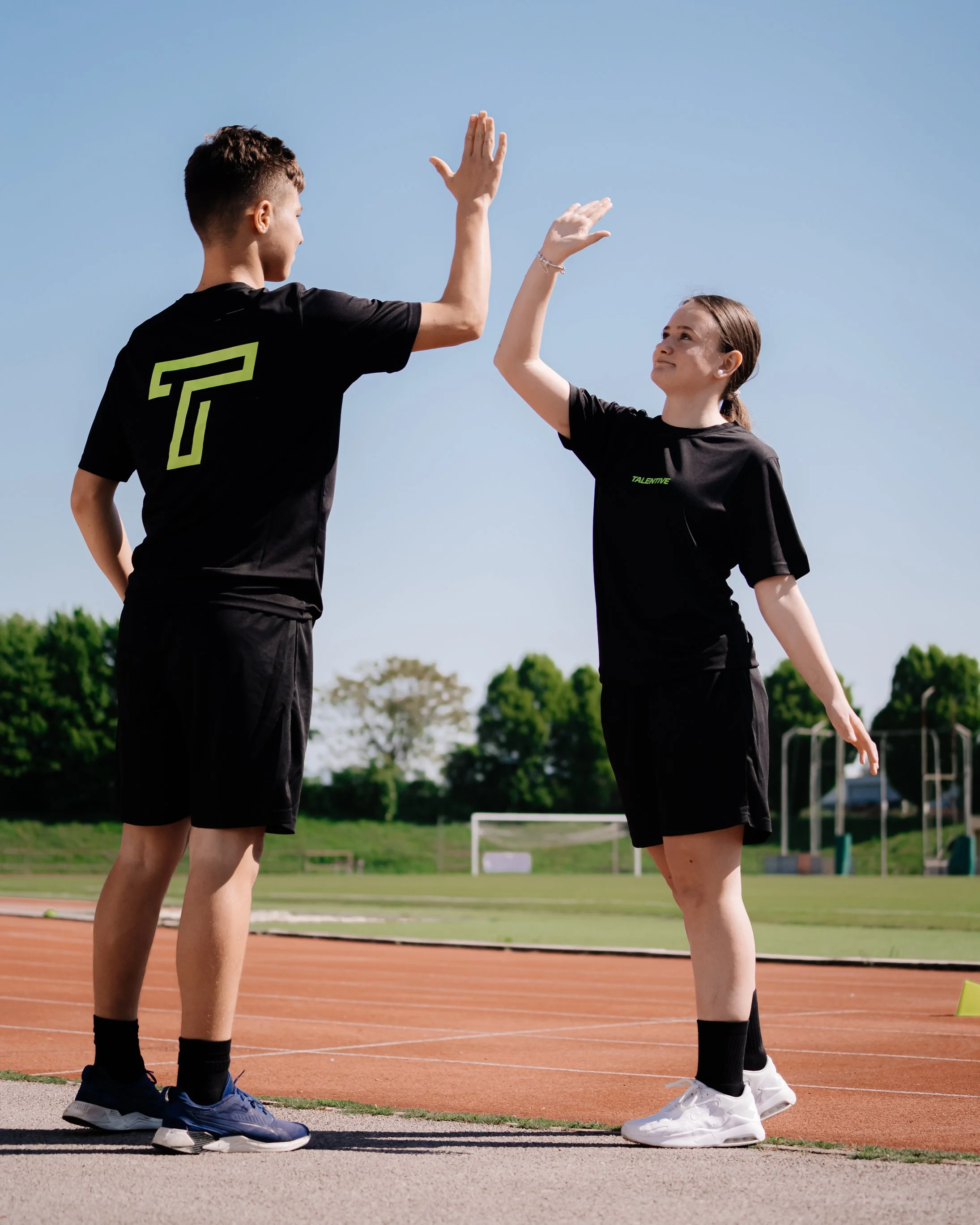 A young male and female athlete giving each other a high five on a running track outdoors on a sunny day.