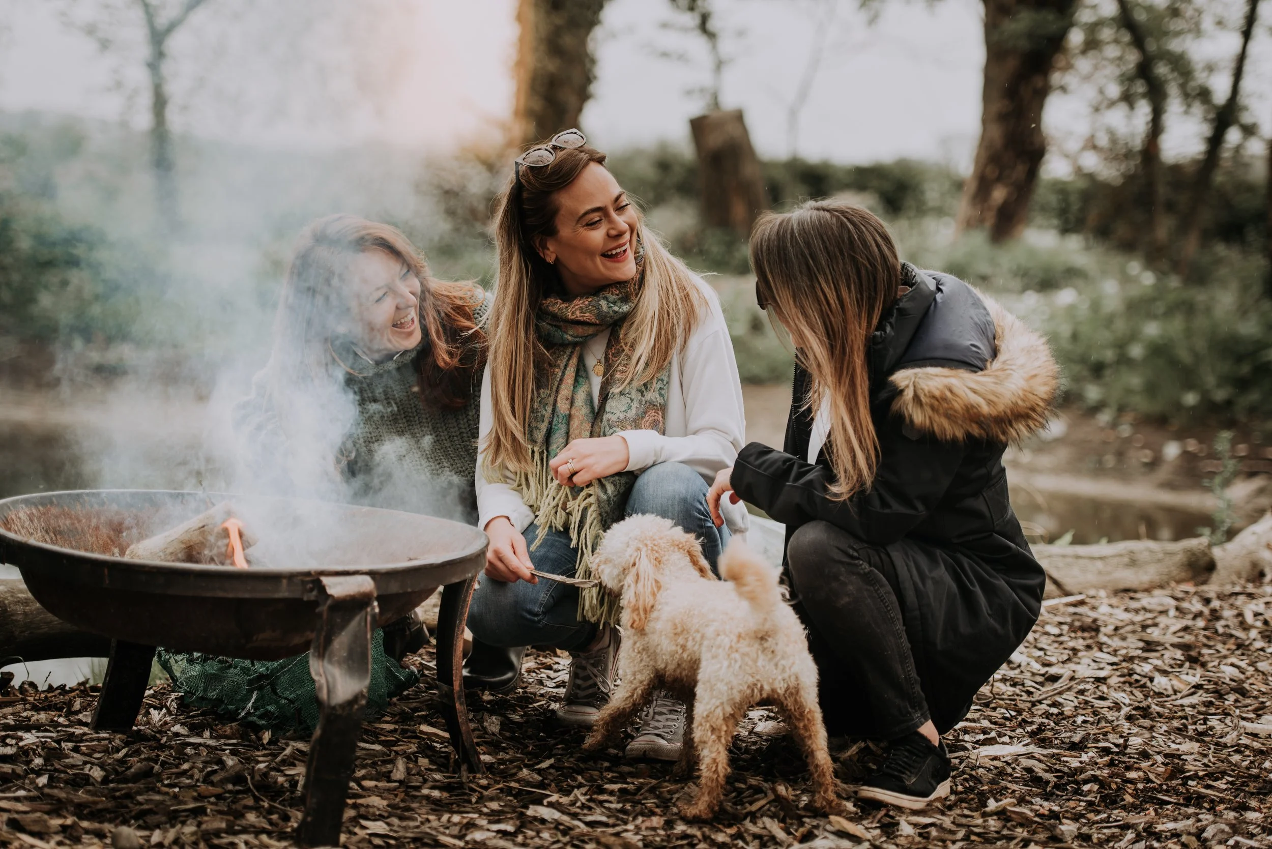 A group of women and a dog are sat by a firepit, all the women are laughing and looking towards each other.