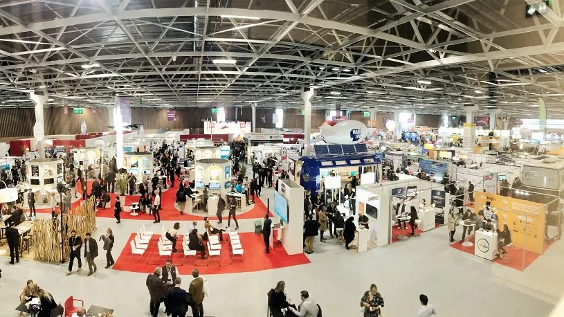 Une exposition ou une foire avec de nombreux stands, visiteurs et zones d'attente dans un grand espace intérieur avec une structure métallique au plafond.
