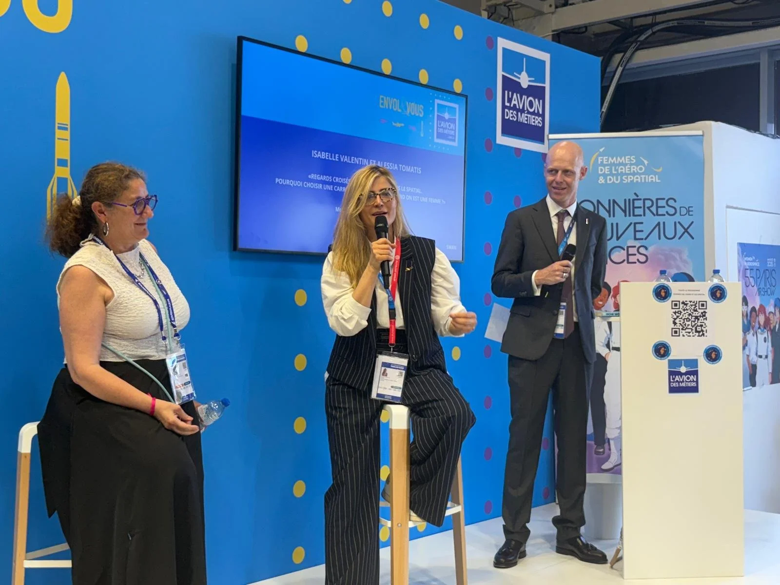 Trois personnes parlent lors d'une conférence avec un décor bleu et des affiches en français, deux hommes et une femme. La femme est assise sur un tabouret, tenant une bouteille d'eau, et parle dans un microphone. Un homme en costume gris et une femme en veston rayé sont aussi présents, avec un écran derrière eux.