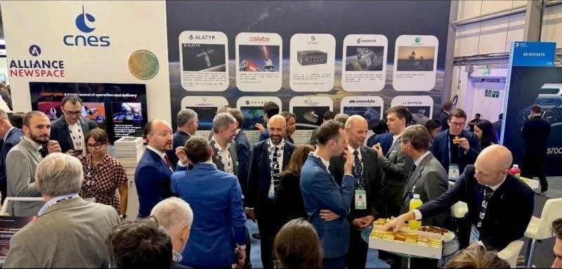 Un groupe de personnes en costume lors d'une conférence ou exposition, avec un stand présentant des logos de sociétés technologiques et un écran affichant des informations.