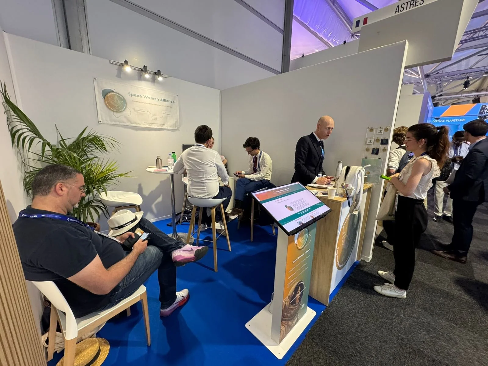 Une scène dans un salon professionnel avec des personnes discutant devant un stand de l'ingénierie spatiale intitulé 'Space Women Alliance'. Un panneau d'information numérique, un stand avec brochures, et plusieurs participants portant des badges. L'ambiance est celle d'une exposition ou d'une conférence.