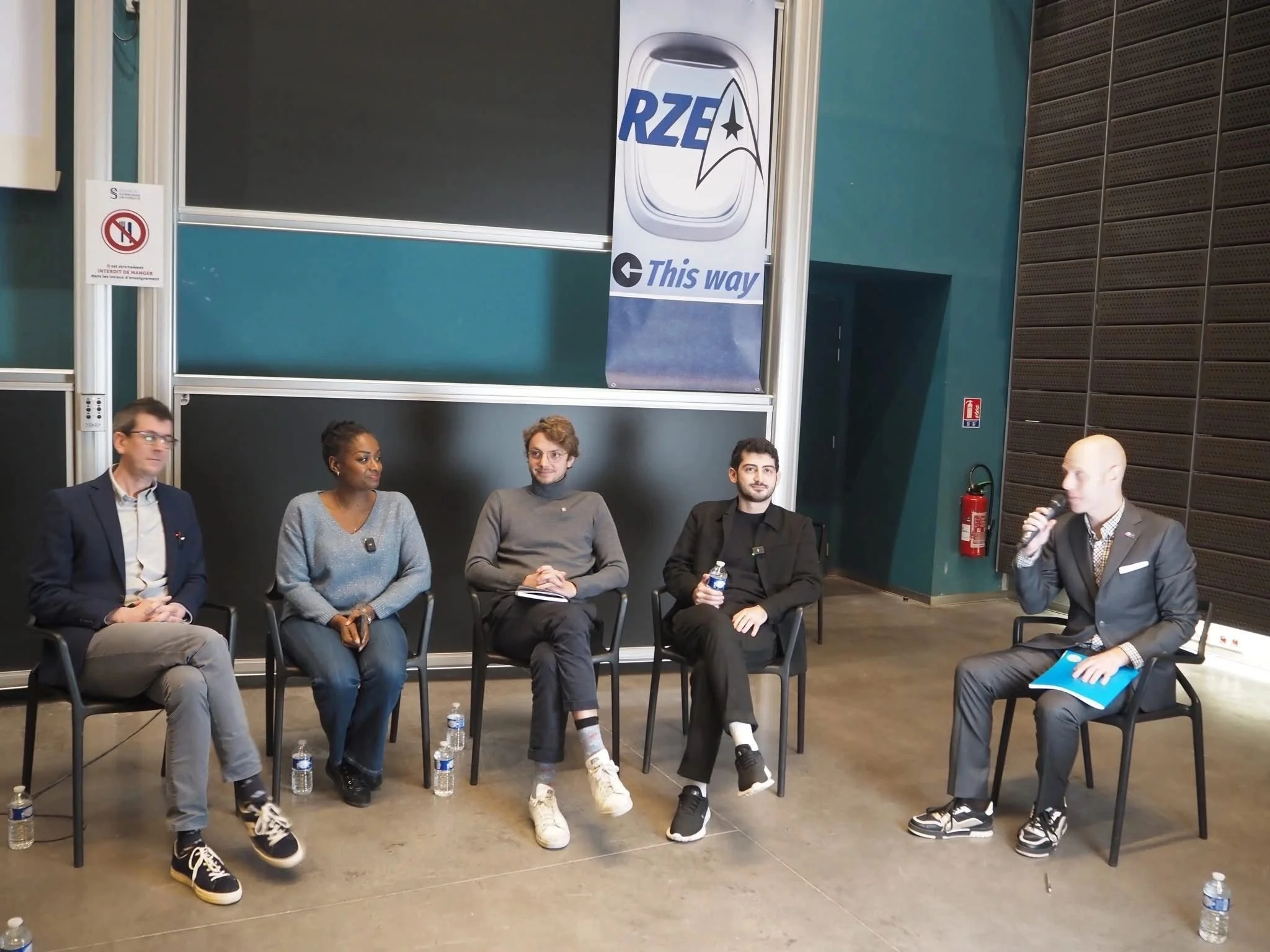 Cinq personnes assises sur des chaises lors d'une discussion ou d'une table ronde, avec un intervenant parlant en micro, dans un espace intérieur avec murs en métal et panneau d'affichage en arrière-plan.