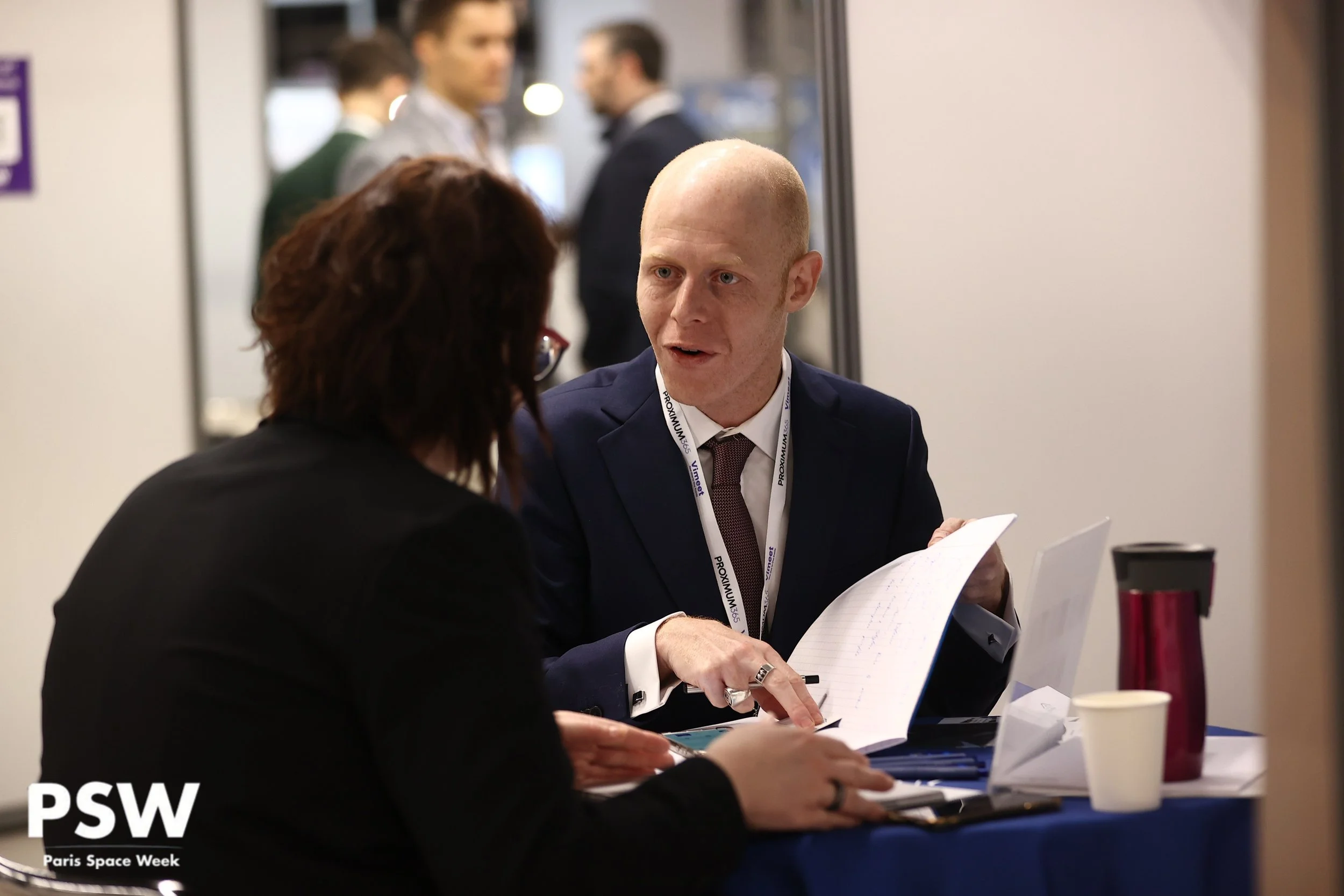 Deux personnes discutent lors de Paris Space Week, assises à une table avec documents, tasses et une thermos. En arrière-plan, d'autres participants échangent.