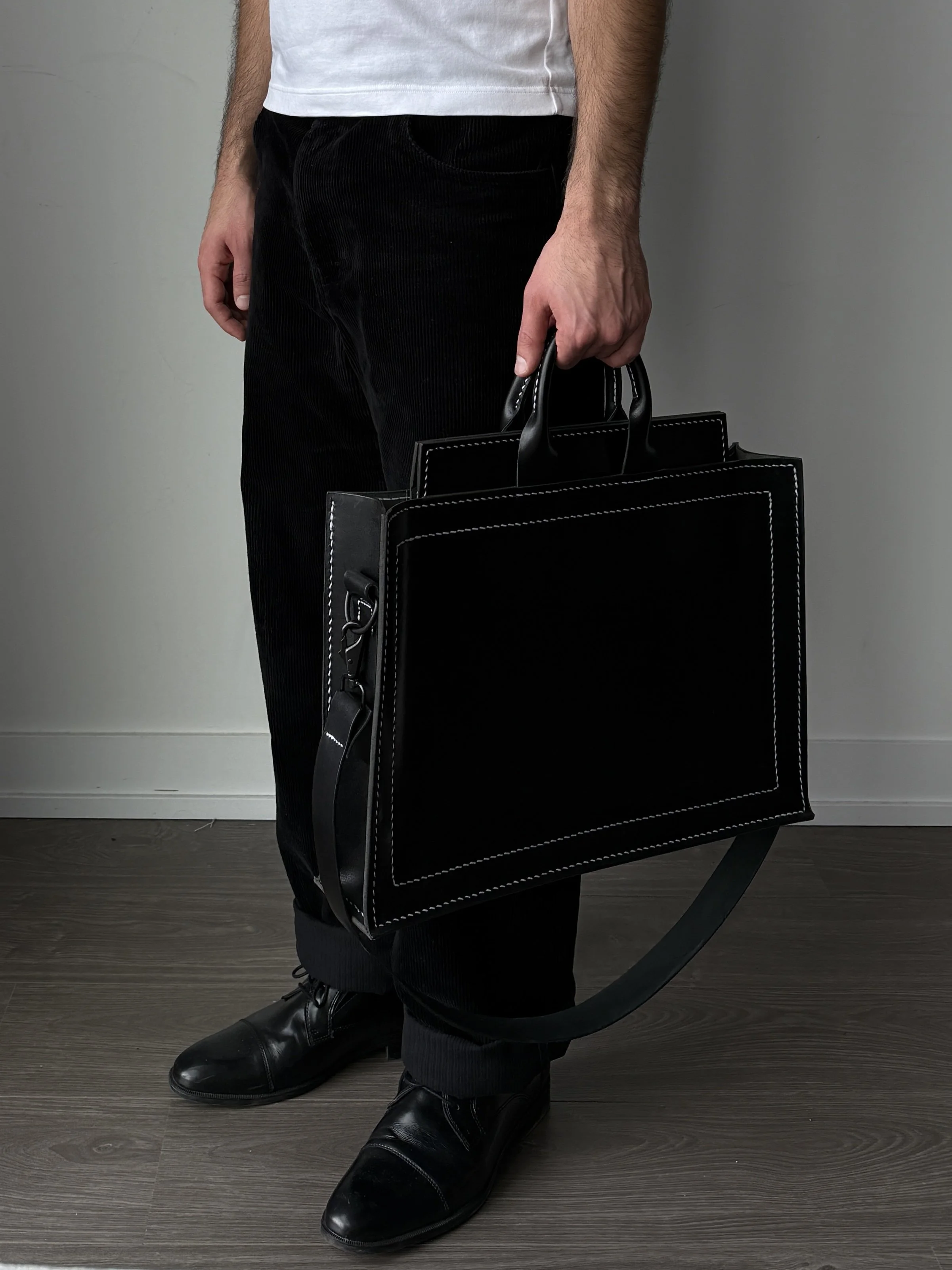 A person dressed in black shoes and black pants, holding a large black bag with white stitching, standing on a dark wooden floor against a plain wall.