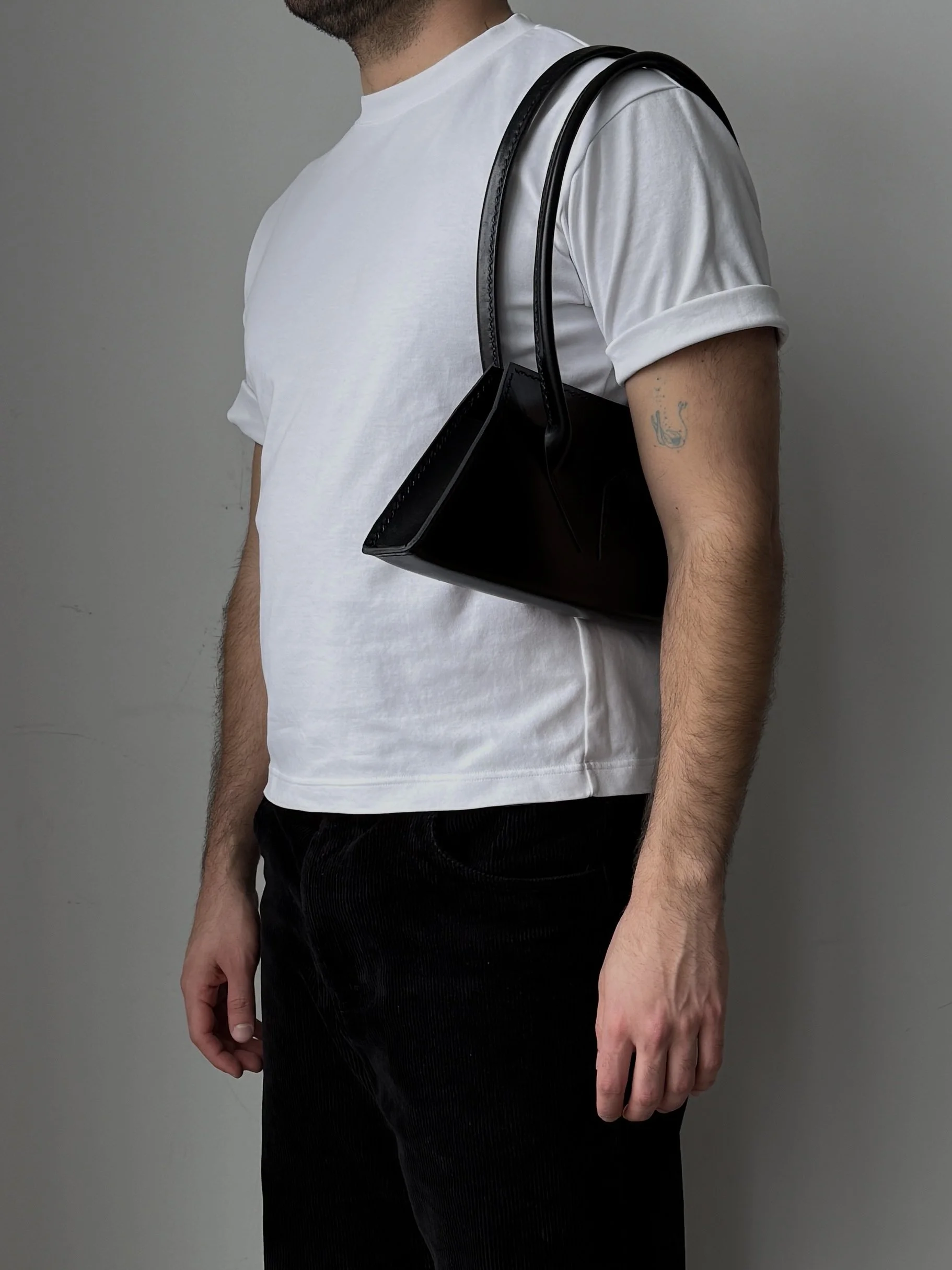 A man wearing a white t-shirt and black pants carrying a black purse on his shoulder.
