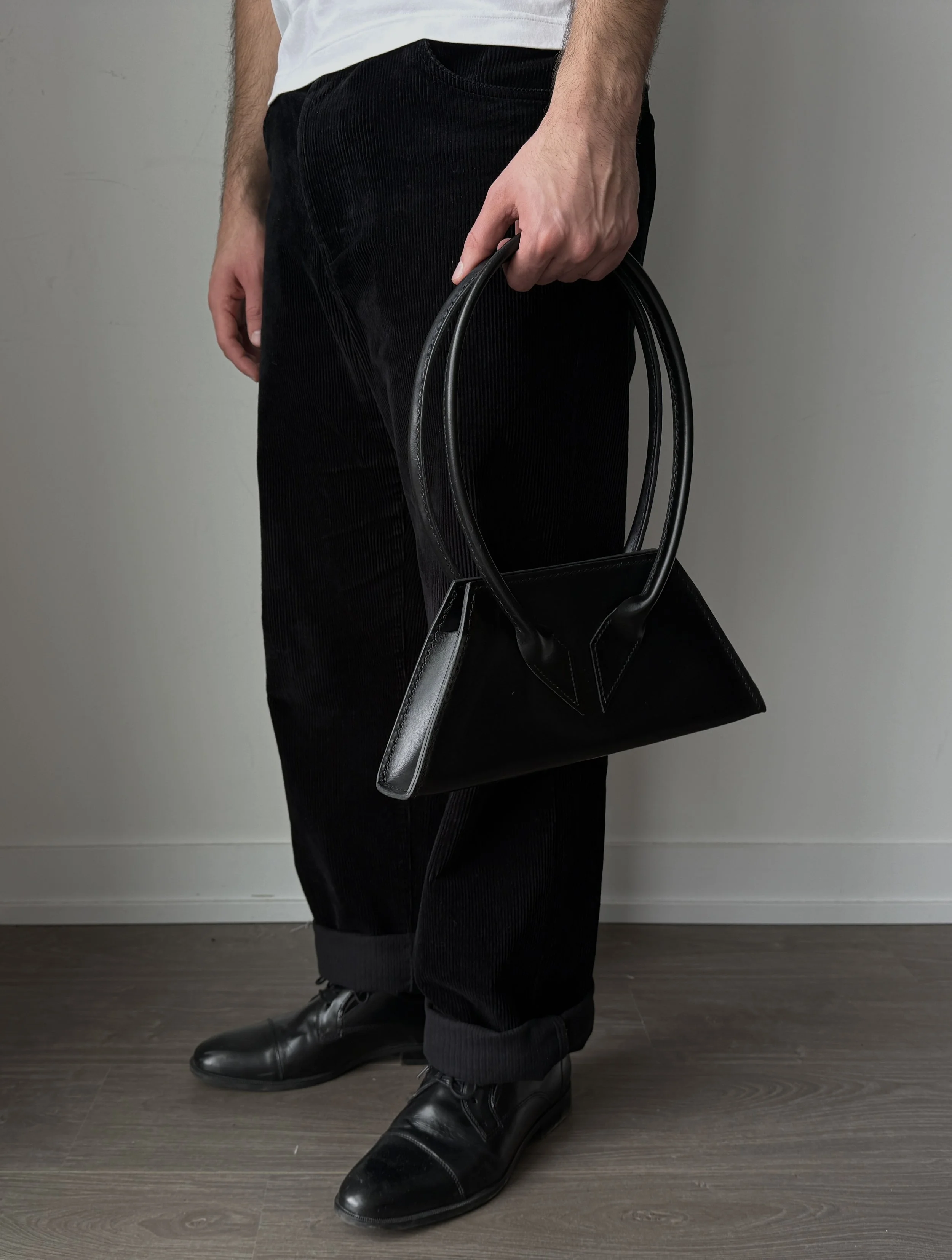 A person wearing black pants and black shoes, holding a black handbag in their right hand, standing on a dark wooden floor against a gray wall.