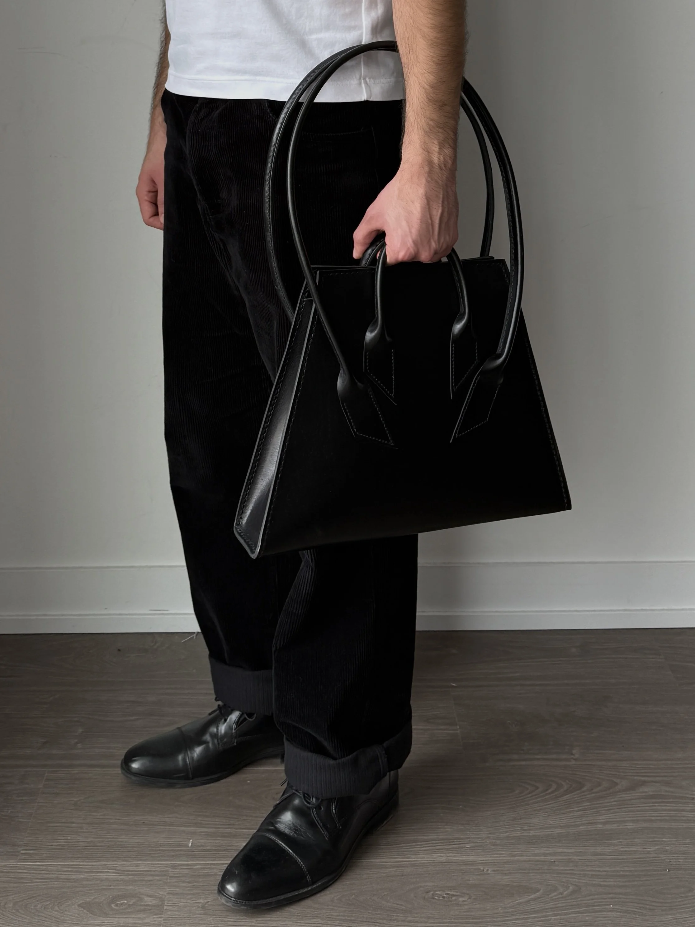 A person dressed in black shoes, black corduroy pants, a white shirt, holding a large black handbag with short handles and a structured shape, standing against a neutral background.